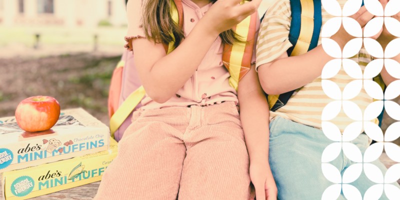 Two young children wearing backpacks sit side-by-side outdoors. Next to them on a wooden bench is a stacked box of Abe&rsquo;s Chocolate Chip Mini Muffins and Corn Mini Muffins with a red apple resting on top. A white geometric pattern runs down the right border of the image.