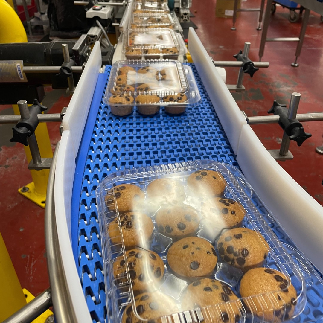 Packages of Abe's vegan chocolate chip mini muffins in clear plastic containers move down a factory assembly line on a blue textured conveyor belt.