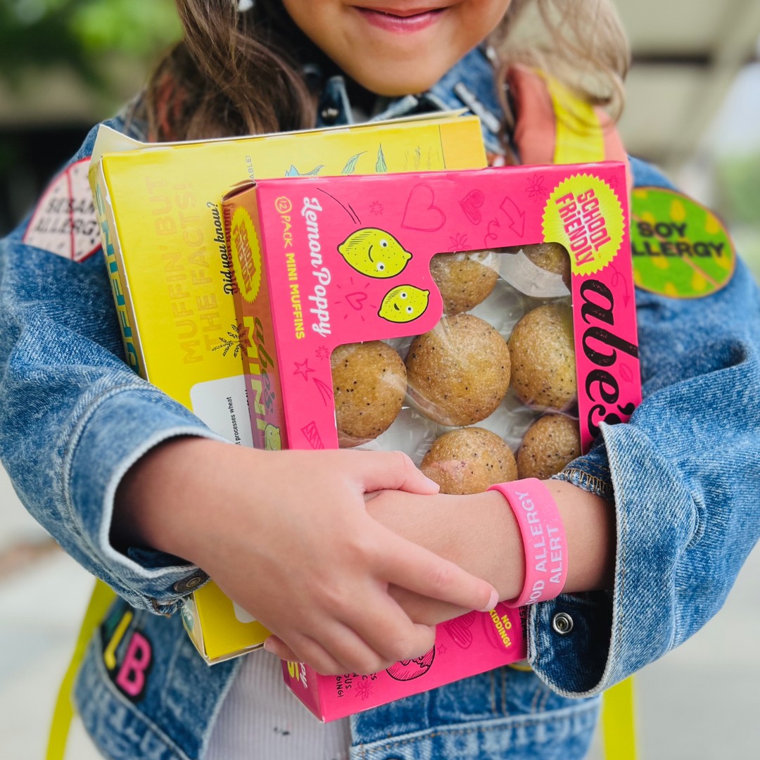 A child wearing a denim jacket covered in allergy awareness patches and a pink "FOOD ALLERGY ALERT" wristband holds tightly onto a pink box of Abe&rsquo;s Lemon Poppy Mini Muffins and a yellow box.