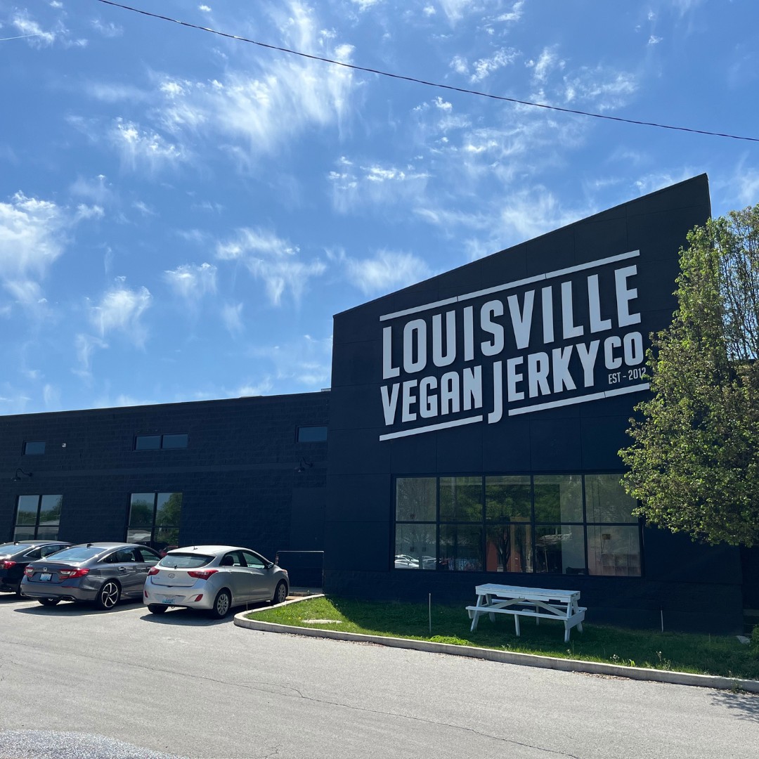 A wide exterior view of the Louisville Vegan Jerky Co. facility. The modern black building features a white painted brand logo on its angled facade, with a parking lot and a white picnic bench on the grass under a bright blue, wispy-cloud sky.