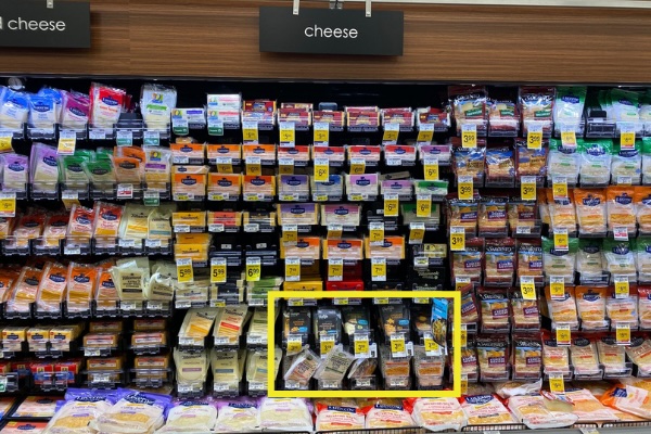 A grocery store refrigerated shelf display stocked with various brands and types of pre-packaged dairy cheeses under a wood-paneled wall sign reading "cheese". A yellow outline box on the lower shelf highlights an integrated section of plant-based cheese products, including Violife slices and shreds, placed directly alongside conventional cheese options.