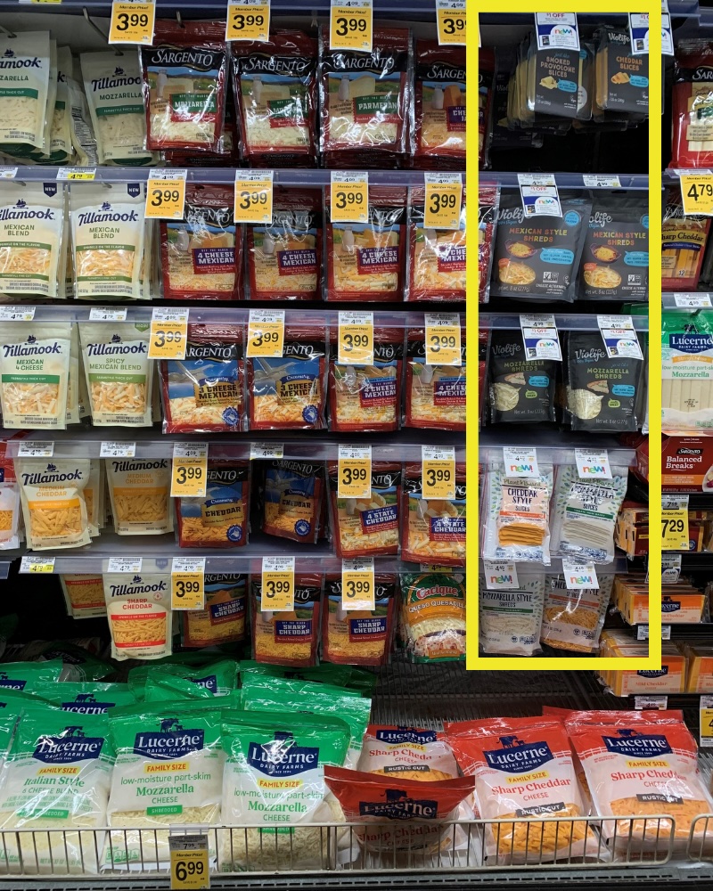 A full-front view of multi-tiered grocery refrigerator shelves organized with bags of shredded and sliced cheeses from brands like Tillamook, Sargento, and Lucerne. A vertical yellow highlight box outlines a column in the middle-right section stocked entirely with plant-based cheeses, including Violife and Open Nature shredded and sliced varieties.