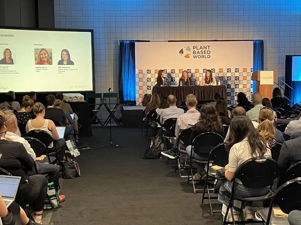 An auditorium viewpoint looking at an event panel on stage at a "PLANT BASED WORLD" convention. Three women sit behind a long draped presentation table speaking into microphones, facing rows of seated audience members. A large projection screen on the far left displays the panel discussion details and headshots of the speakers.