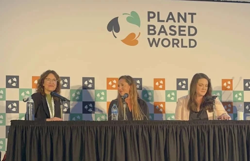 A close-up view of three women participating in a panel discussion, seated behind a black fabric-draped table. The stage backdrop features a patterned design of green, white, blue, and orange squares underneath a large logo that reads "PLANT BASED WORLD".