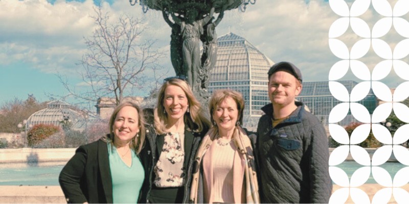 Four smiling professionals stand shoulder-to-shoulder outside in front of a stone fountain. A large glass conservatory building is visible in the background under a blue sky, and a white geometric pattern frames the right edge of the image.
