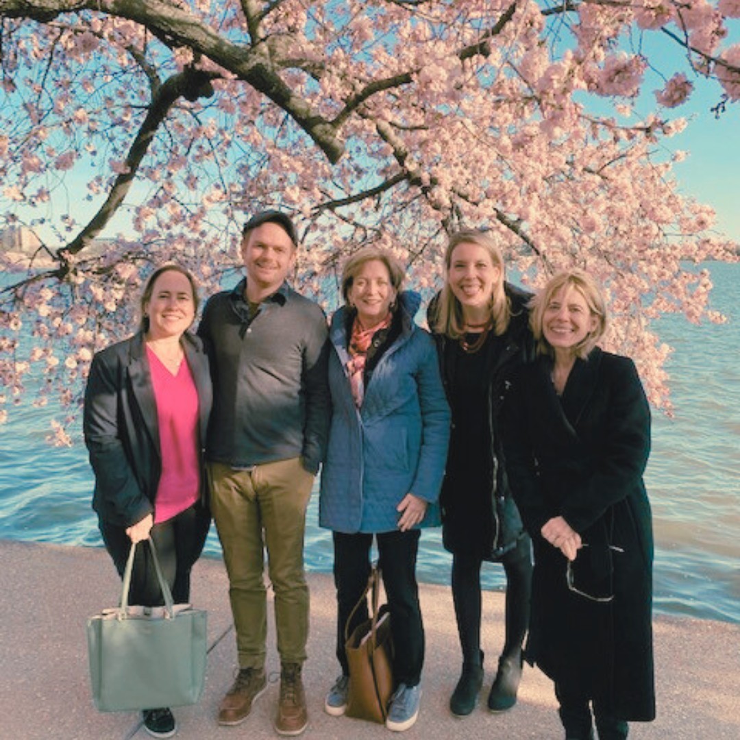 Five smiling people stand in a row along a paved walkway directly beneath a large cherry blossom tree in full pink bloom. A body of water stretches across the background.