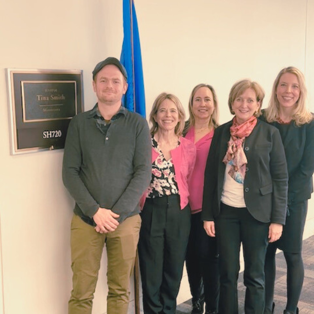 Five colleagues pose for a group photo in an office hallway next to a wall plaque that reads "SENATOR Tina Smith, Minnesota, SH720". A blue flag on a pole stands right behind them.