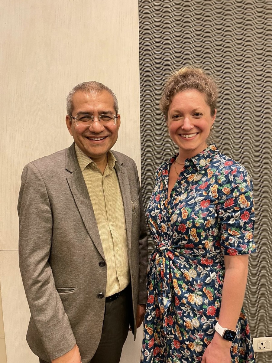 A man wearing glasses and a grey blazer stands side-by-side smiling next to a woman wearing a short-sleeved, colorful floral print dress against a patterned textured wall.