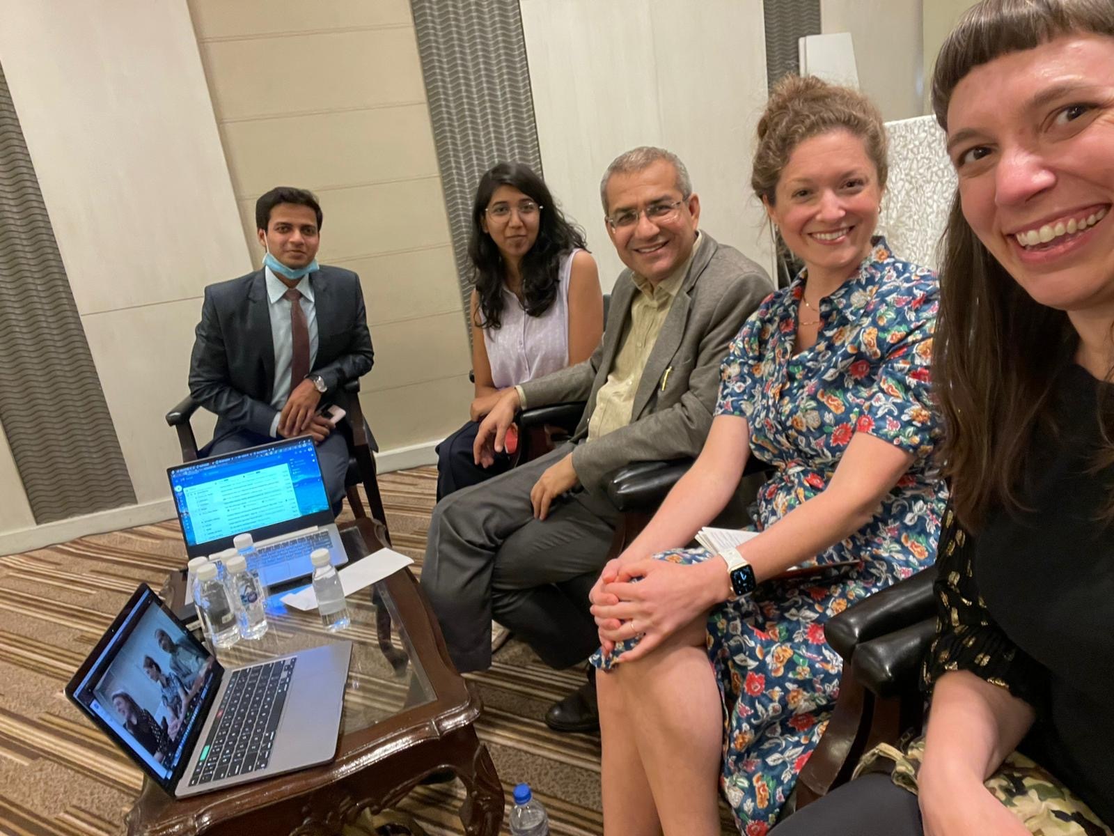 A group of five professionals participating in a hybrid meeting inside a conference room. A woman takes a close-up selfie showing colleagues seated together in armchairs next to a coffee table with water bottles and two open laptops, one displaying a live video conference call.