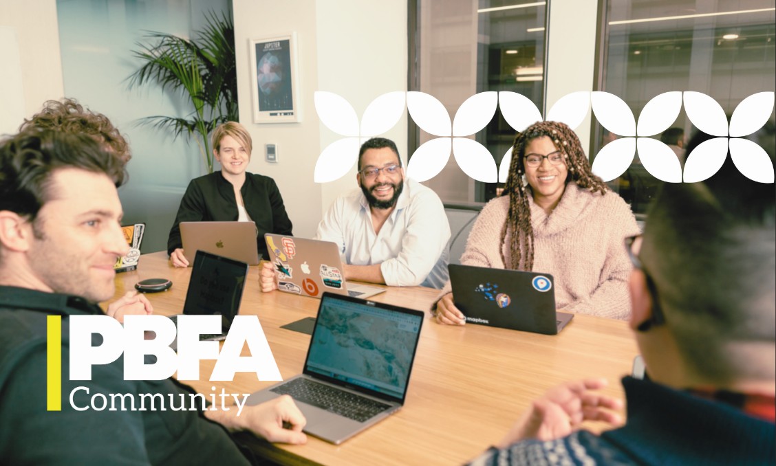A group of five professionals sit around a light wooden conference table during a meeting, smiling and working on their open laptops. A logo in the bottom-left corner reads "PBFA Community" beneath a yellow accent bar, and a white geometric flower pattern stretches across the top right.