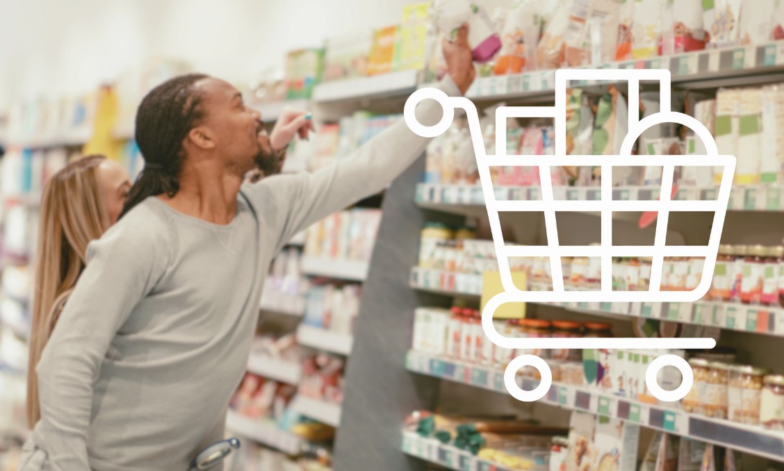 A man with long dark hair tied back reaches for an item on a high grocery store shelf while shopping with a companion. A large white shopping cart graphic filled with boxes is overlaid on the right side of the image.
