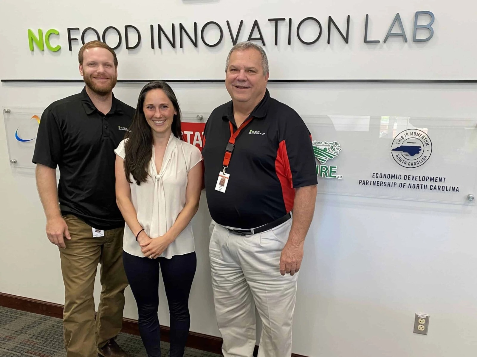 Three smiling professionals stand together in front of a clean white wall displaying large text that reads "NC FOOD INNOVATION LAB". Behind them, a clear acrylic panel is mounted to the wall, showcasing various organizational logos, including the "Economic Development Partnership of North Carolina."
