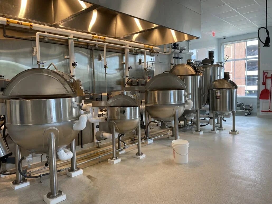An industrial pilot plant kitchen featuring a row of large, stainless-steel hemispherical cooking kettles with their lids partially open. The machinery is connected by a system of overhead utility piping beneath heavy-duty ventilation hoods.