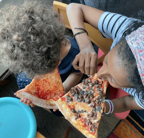 An overhead shot of a woman with her hair in a headband and a young child with curly hair sitting side-by-side, each taking a bite out of their own large slice of pizza. The woman's slice is heavily topped with chopped black olives.