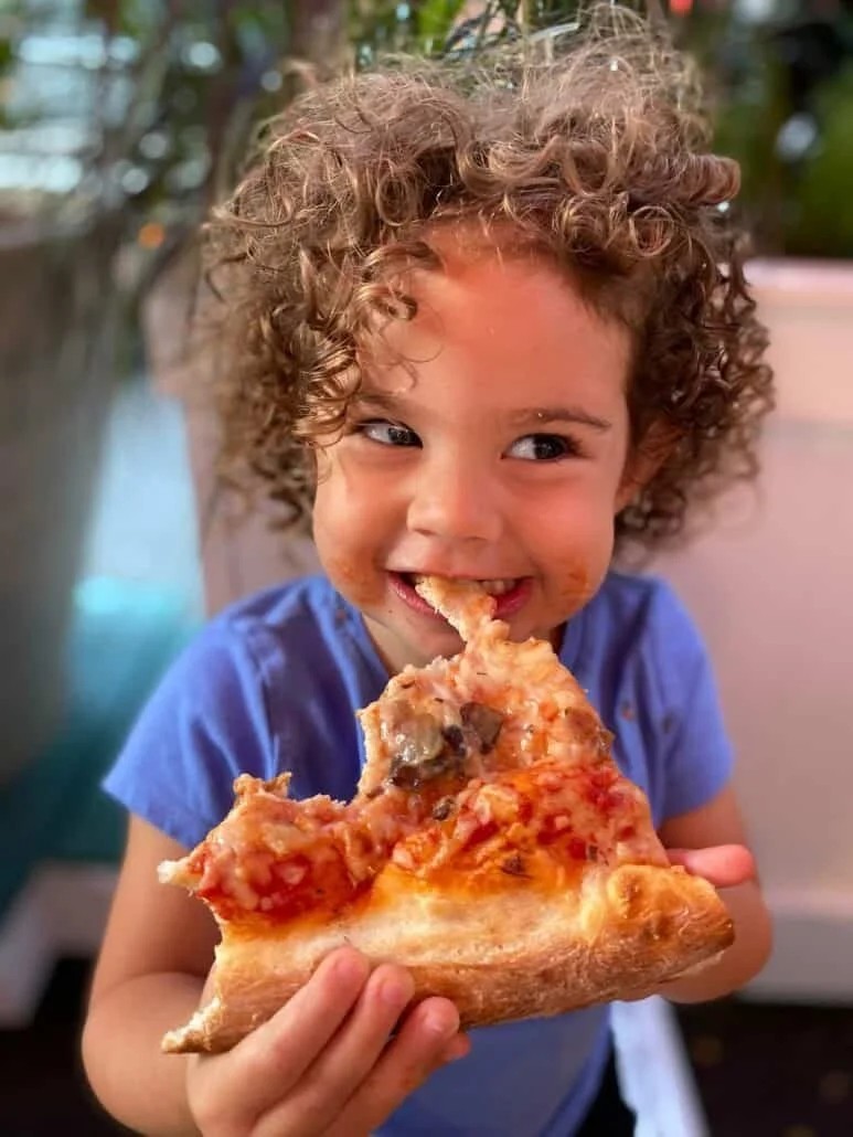 A close-up portrait of a smiling young child with tight brown curls wearing a blue t-shirt, happily eating a large, thick-crust slice of cheese pizza with tomato sauce on their face.