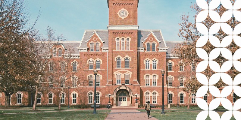 A large, historic brick university building with a central tower and clock dominates the scene. A lone person walks on a path, surrounded by trees with autumn leaves.