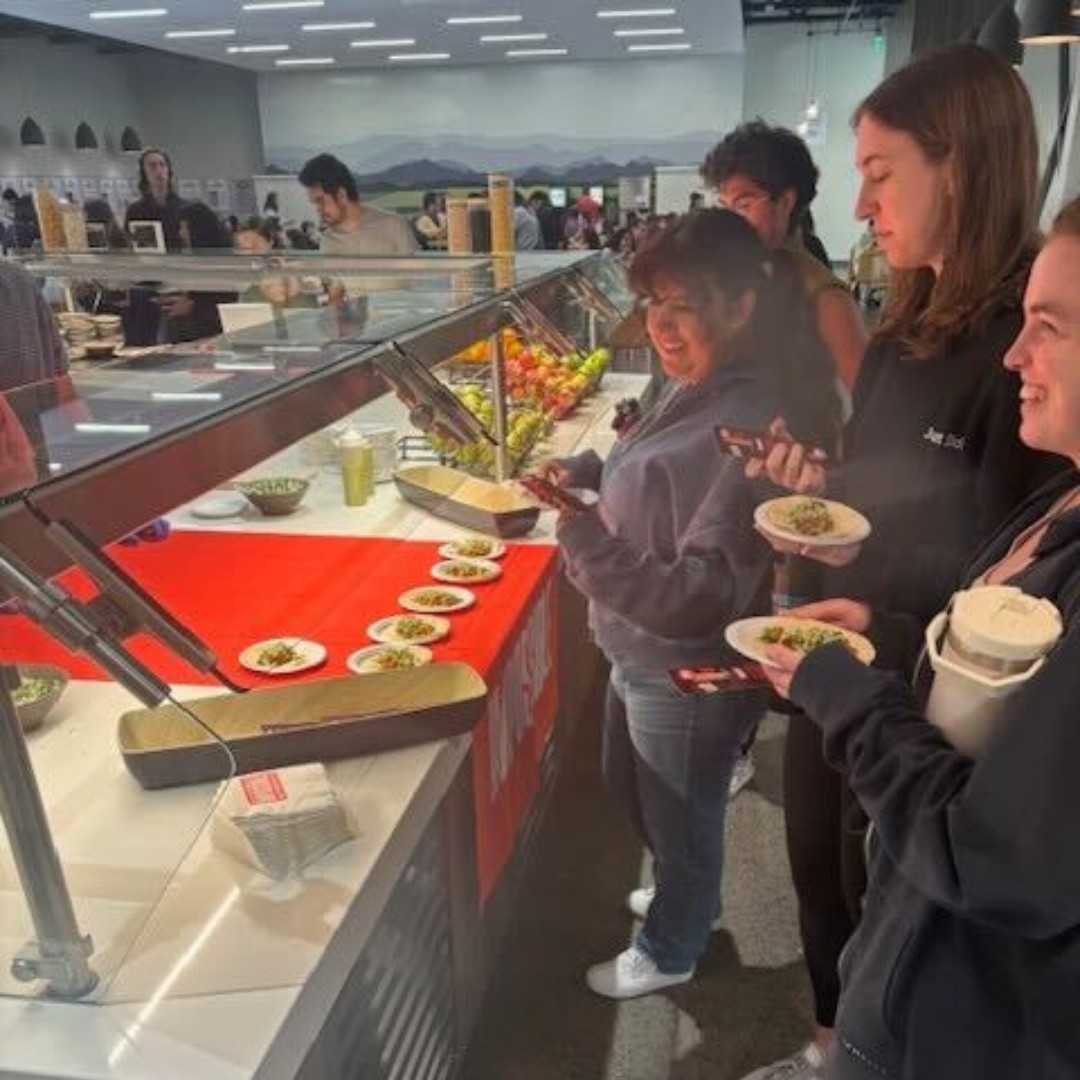People in line at a food counter selecting small plates of food in a brightly lit cafeteria or buffet setting.