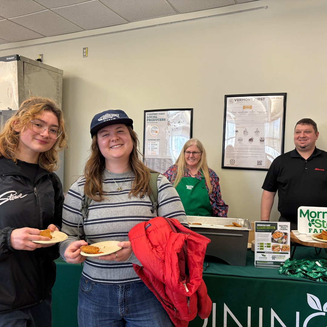 Two students smiling and holding paper plates with small food samples in front of a dining service station. Behind the green-clothed table, two university dining staff members smile at the camera. Informational posters about local producers and dining metrics hang on the wall behind them.
