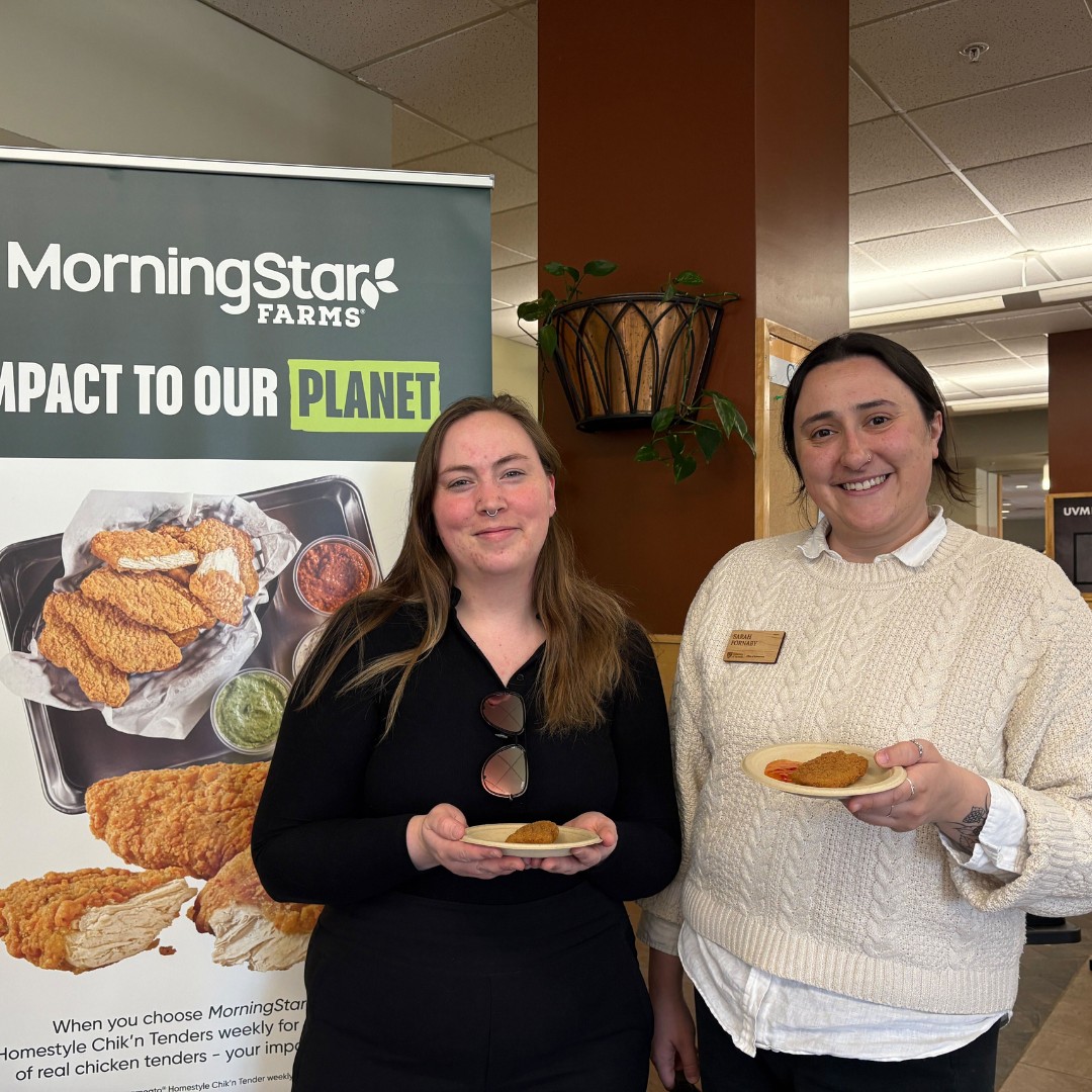 Two women standing side-by-side, smiling and holding small paper plates with plant-based chicken nuggets. They are standing next to a large vertical banner that reads "MorningStar Farms: Impact to Our Planet" featuring a picture of crispy chick'n tenders.