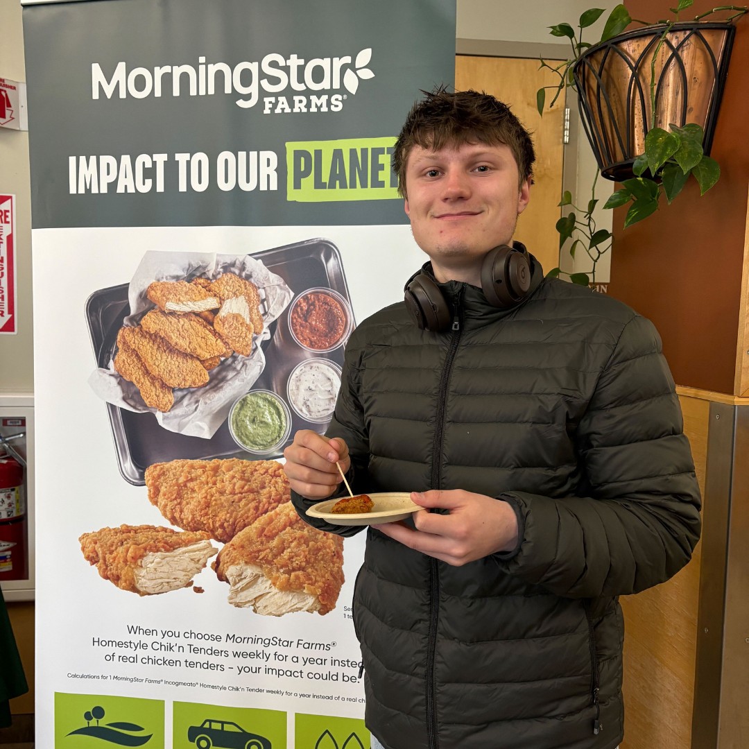 A young man wearing a black puffer jacket and headphones around his neck smiles while holding a small paper plate. He is using a toothpick to pick up a food sample. He stands in front of a large MorningStar Farms promotional banner about the positive environmental impact of plant-based food.