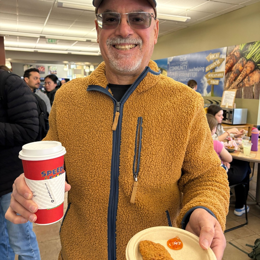 A close-up shot of a smiling man with glasses and a goatee, wearing a textured brown fleece jacket. He holds a red disposable coffee cup in one hand and a paper plate with a plant-based nugget and dipping sauce in the other, inside a busy campus dining hall.