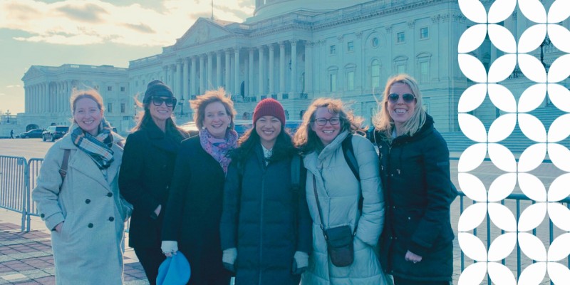PBFA members and staff bundled in winter coats and scarves stand in a row, smiling together on the plaza outside the United States Capitol building on a bright day. A white geometric pattern frames the right edge of the image.