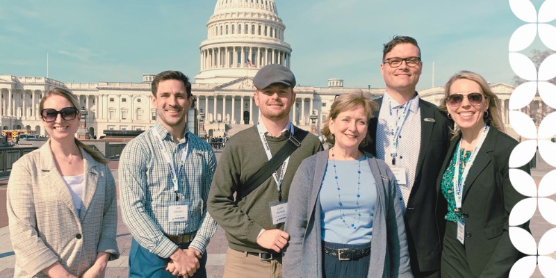 Six smiling professionals stand together in a row outdoors with the United States Capitol dome rising directly behind them. They are wearing credentials on lanyards, and a white geometric pattern frames the right edge of the image.