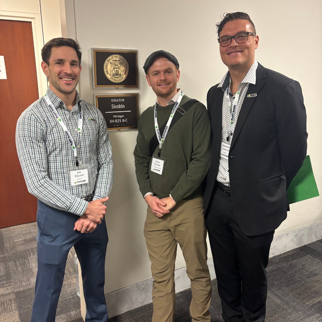 Three men wearing conference lanyards stand and smile in an office hallway next to a dark wall plaque reading "SENATOR Slotkin, Michigan, SH-825 B-C".