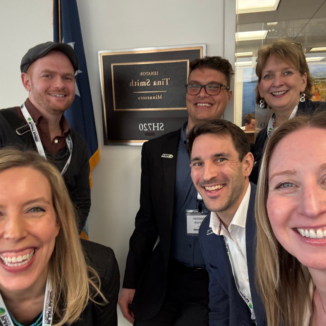 A close-up selfie of six smiling people gathered together in a government office. An official plaque on the wall behind them reads "SENATOR Tina Smith, Minnesota, SH720" in mirrored text, with an American flag visible on the left.