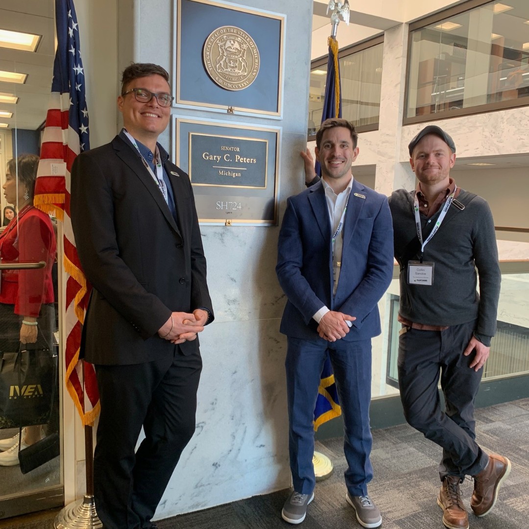 Three professionals stand in a brightly lit office corridor next to an American flag and a gold wall sign reading "SENATOR Gary C. Peters, Michigan, SH724".