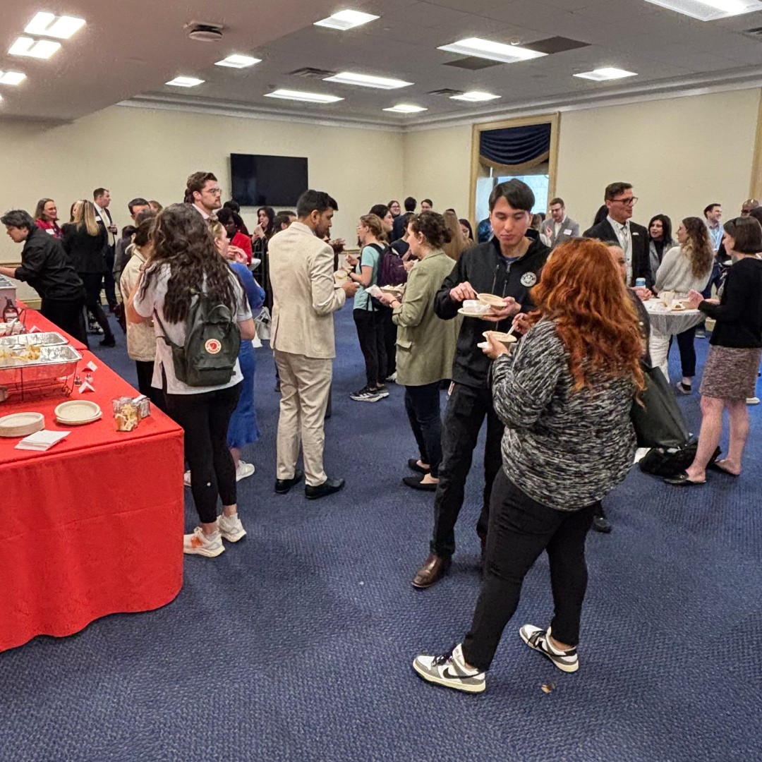 A busy indoor reception hall where numerous attendees are standing, talking, and eating from small paper plates. A long table draped in a bright red tablecloth on the left holds catering trays and additional plates.