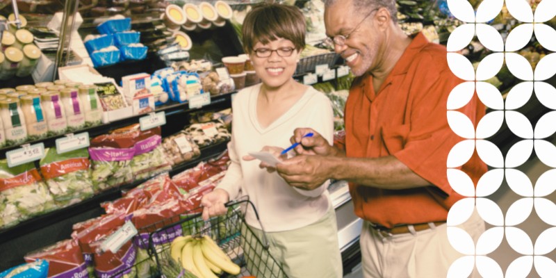 A smiling older man and woman looking over a shopping list together in the produce section of a grocery store. The woman holds a shopping cart handle, while the man points a pen at the paper list. A white geometric pattern runs down the right border of the image.