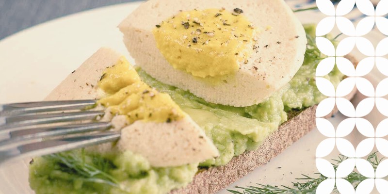 A slice of toast topped with mashed green avocado and a sliced plant-based hard-boiled egg substitute, seasoned with black pepper and fresh dill. A fork presses into one of the egg pieces. A white geometric pattern frames the right edge of the image.