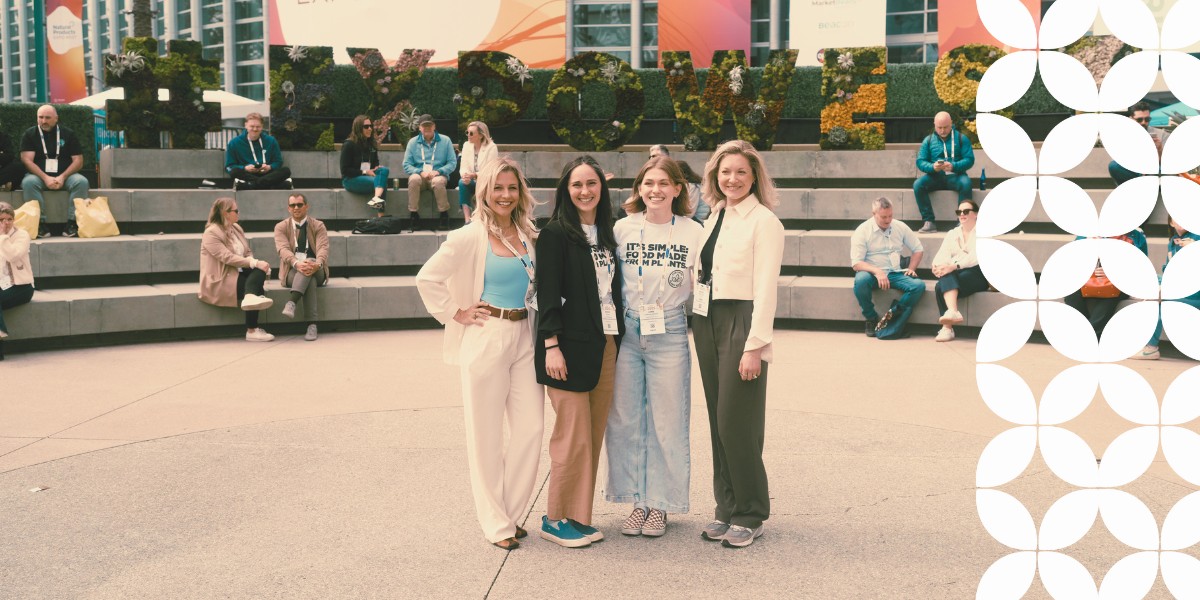 Four women smiling and standing together on an open-air plaza. In the background, tiered concrete seating is filled with event attendees, and a massive green topiary sign reads "#EXPOWEST". A white geometric pattern frames the right edge.
