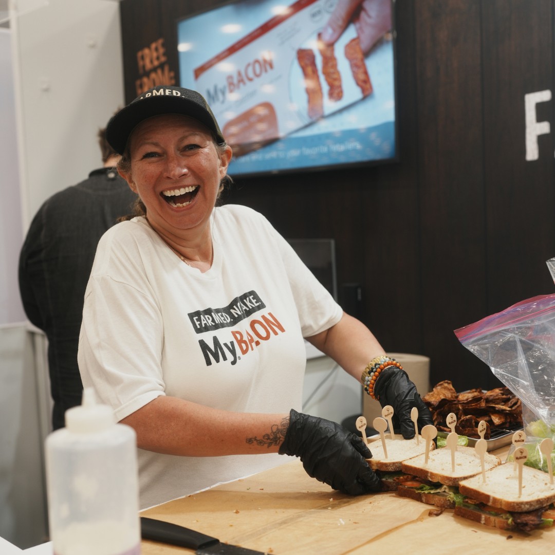 A woman laughing joyfully while wearing a black cap and a white t-shirt that says "Farmed. Nake. MyBacon." Wearing black gloves, she prepares a row of sandwiches on a wooden cutting board, with a digital sign for "MyBacon" behind her.