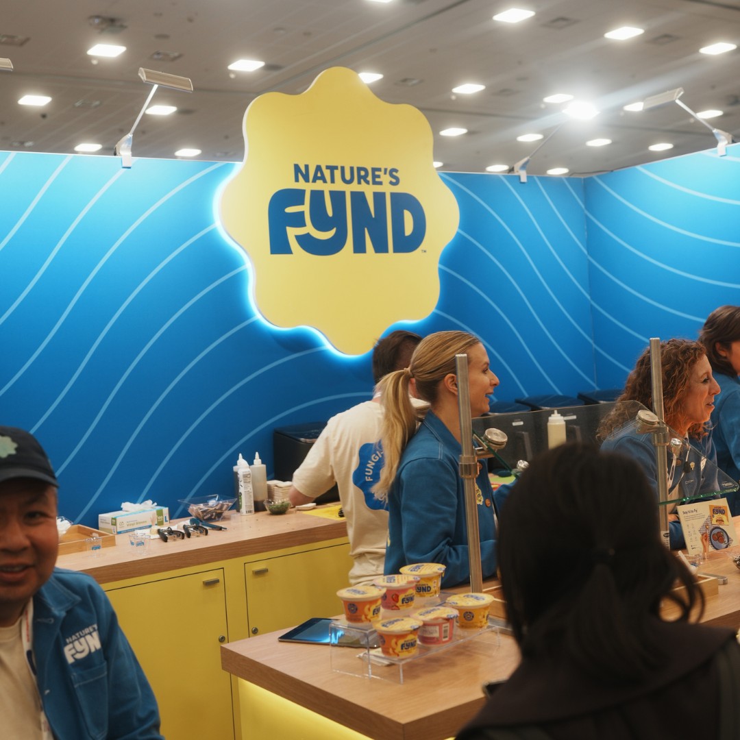 At a trade show booth, staff members in blue shirts stand behind a yellow counter beneath a large yellow flower-shaped sign that reads "NATURE'S FYND". Small sample cups of yogurt are displayed on a clear tray on the counter.