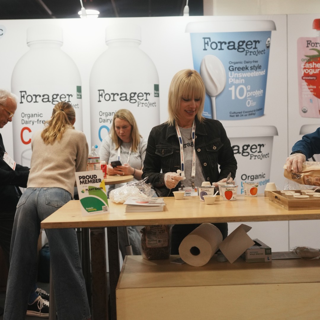 A blonde woman wearing a black denim jacket and plastic gloves prepares food samples at a Forager Project convention booth. Large backdrops behind her display images of organic dairy-free milk bottles and Greek-style unsweetened plain yogurt containers.