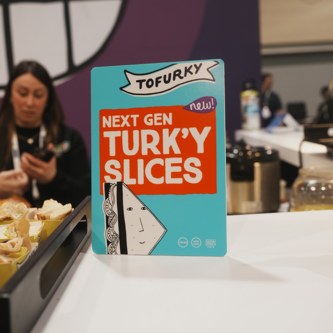 A colorful promotional card on a counter reads "TOFURKY new! NEXT GEN TURK'Y SLICES" with an illustration of a sandwich face. On the left, a tray holds actual sandwich samples, while a blurred woman looks down at her phone in the background.