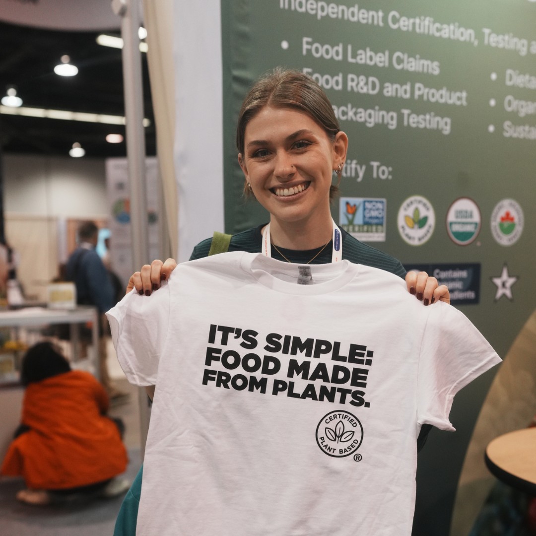A smiling woman holds up a white t-shirt that reads "IT'S SIMPLE: FOOD MADE FROM PLANTS." with a "Certified Plant Based" logo below the text. She is standing in front of a green trade show banner detailing food certification services.