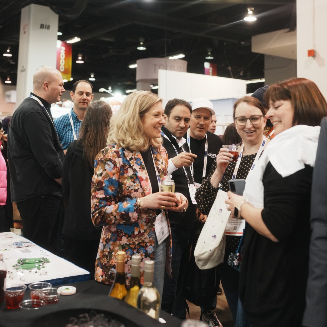 A group of people socializing and networking in a crowded indoor convention hall. A woman in a colorful floral blazer smiles while holding a drink and talking with other attendees near a table.