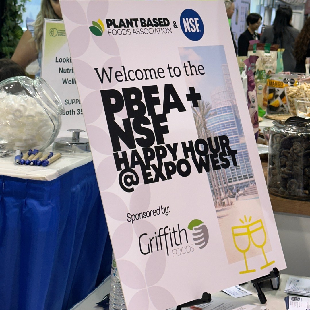 A close-up of a promotional easel sign at a trade show reading "Welcome to the PBFA + NSF HAPPY HOUR @ EXPO WEST, Sponsored by: Griffith Foods". A large clear bowl filled with mints rests on the table nearby.
