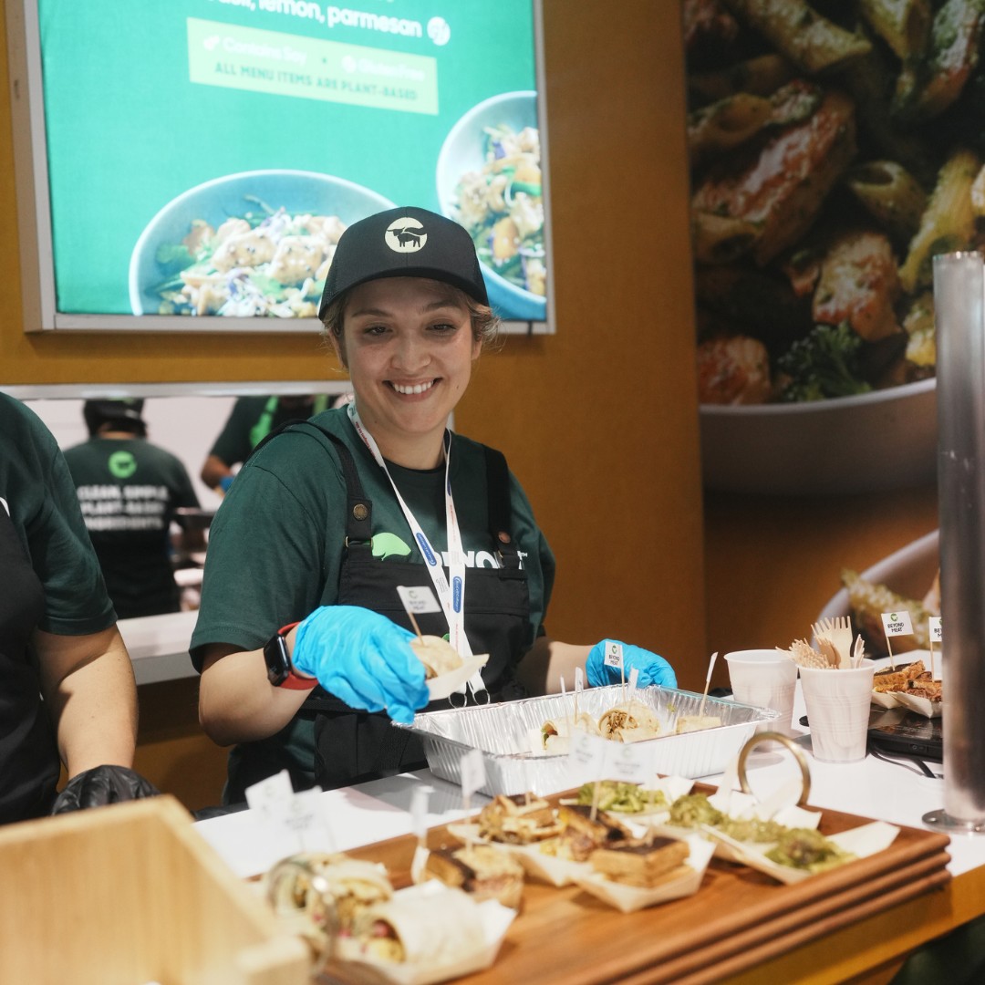 A smiling food vendor wearing a black cap, green t-shirt, and blue disposable gloves serves small plant-based wraps from a metal tray at a sampling counter. Additional plates of food samples are lined up on a wooden board in the foreground.