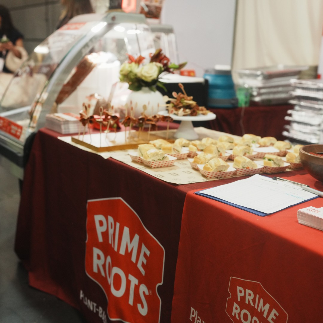 A trade show display table covered in a red tablecloth featuring the "Prime Roots Plant-Based" logo. Trays of miniature sandwiches and toothpick-skewered appetizers are arranged across the table for tasting.