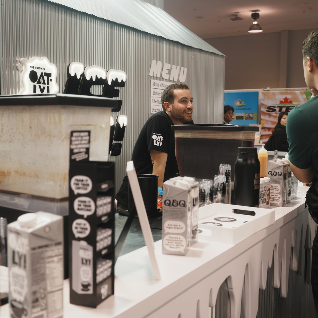 A man wearing an "Oat-ly!" t-shirt stands behind a white serving counter at a conventional convention booth, smiling as he interacts with an attendee. Large beverage dispensers and Oat-ly cartons sit on the counter.