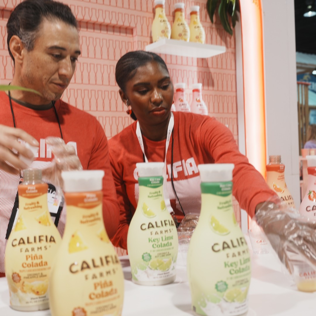 A man and a woman wearing red Califia Farms long-sleeve shirts and plastic gloves pour beverage samples at a white counter. Several bottles of Califia Farms Pi&ntilde;a Colada and Key Lime Colada are displayed in the foreground.