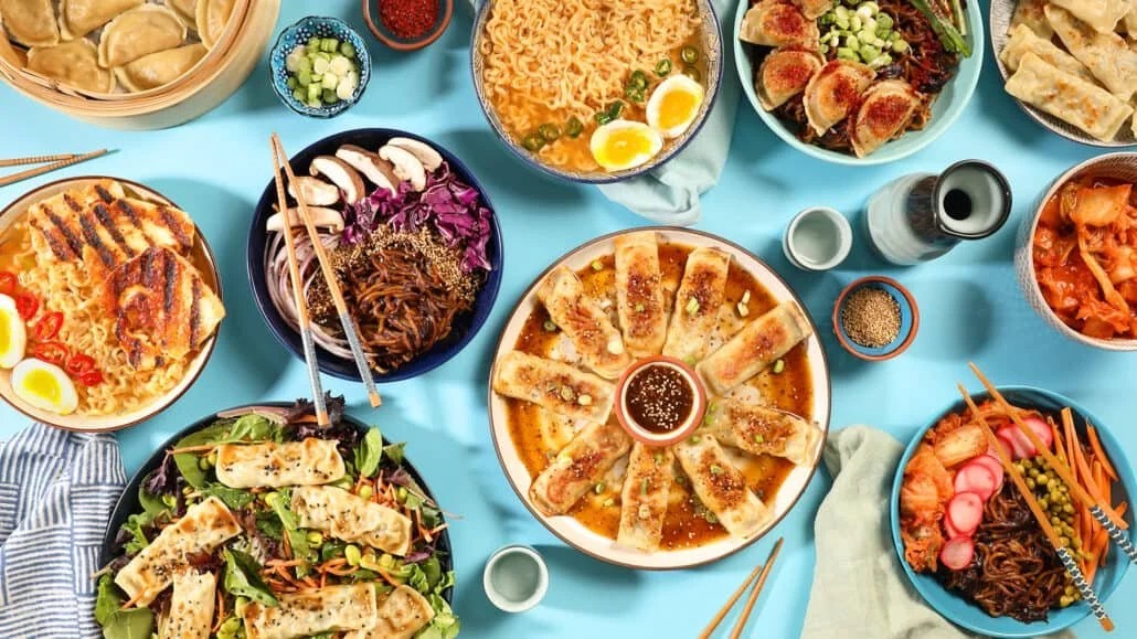 An overhead food flat lay of a diverse Asian feast arranged on a bright blue background. The spread includes various styles of dumplings, ramen bowls with boiled eggs, noodle dishes with sliced mushrooms and purple cabbage, fresh salad, kimchi, small bowls of garnishes, and a central circular plate of pan-fried potstickers surrounding a dipping sauce.