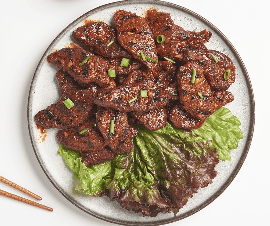A top-down view of a round ceramic plate holding pieces of grilled, marinated plant-based bulgogi meat garnished with chopped green onions. The meat rests on green and purple lettuce leaves, with the tips of wooden chopsticks visible in the bottom left corner against a white background.