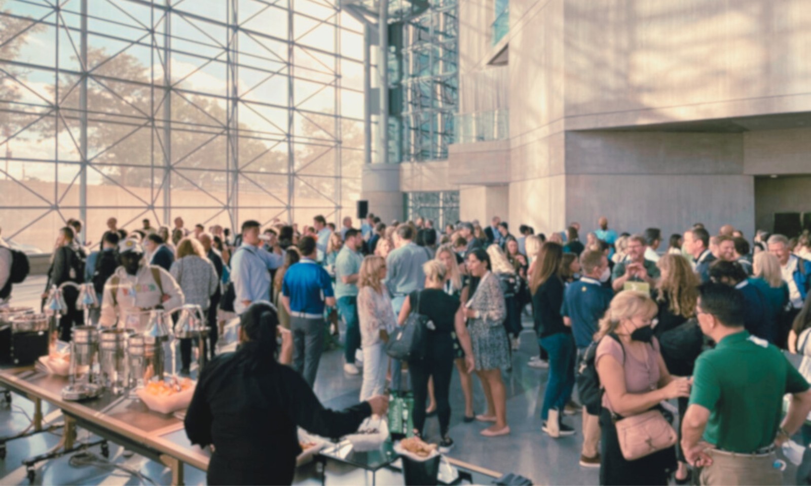 A wide-angle view of a crowded, sunlit networking reception inside a modern building with a massive glass window wall. Attendees stand in groups conversing near a buffet table lined with food trays and serving warmers in the foreground.