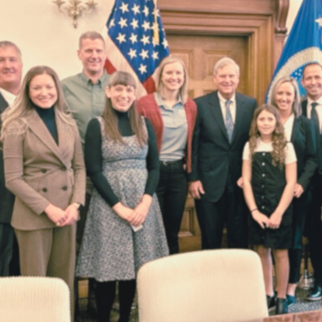 A group photo of several professionals standing together indoors alongside a young girl, posing in front of a wooden wall. A United States flag stands prominent behind them.