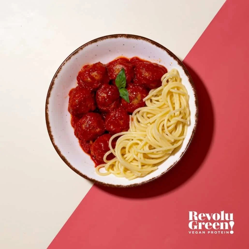 A top-down view of a bowl containing plain spaghetti noodles paired with several plant-based meatballs heavily coated in rich red marinara sauce, garnished with a fresh basil leaf. The bowl rests on a diagonally split cream and red background with a logo in the bottom right corner reading "Revolu Green! VEGAN PROTEIN".
